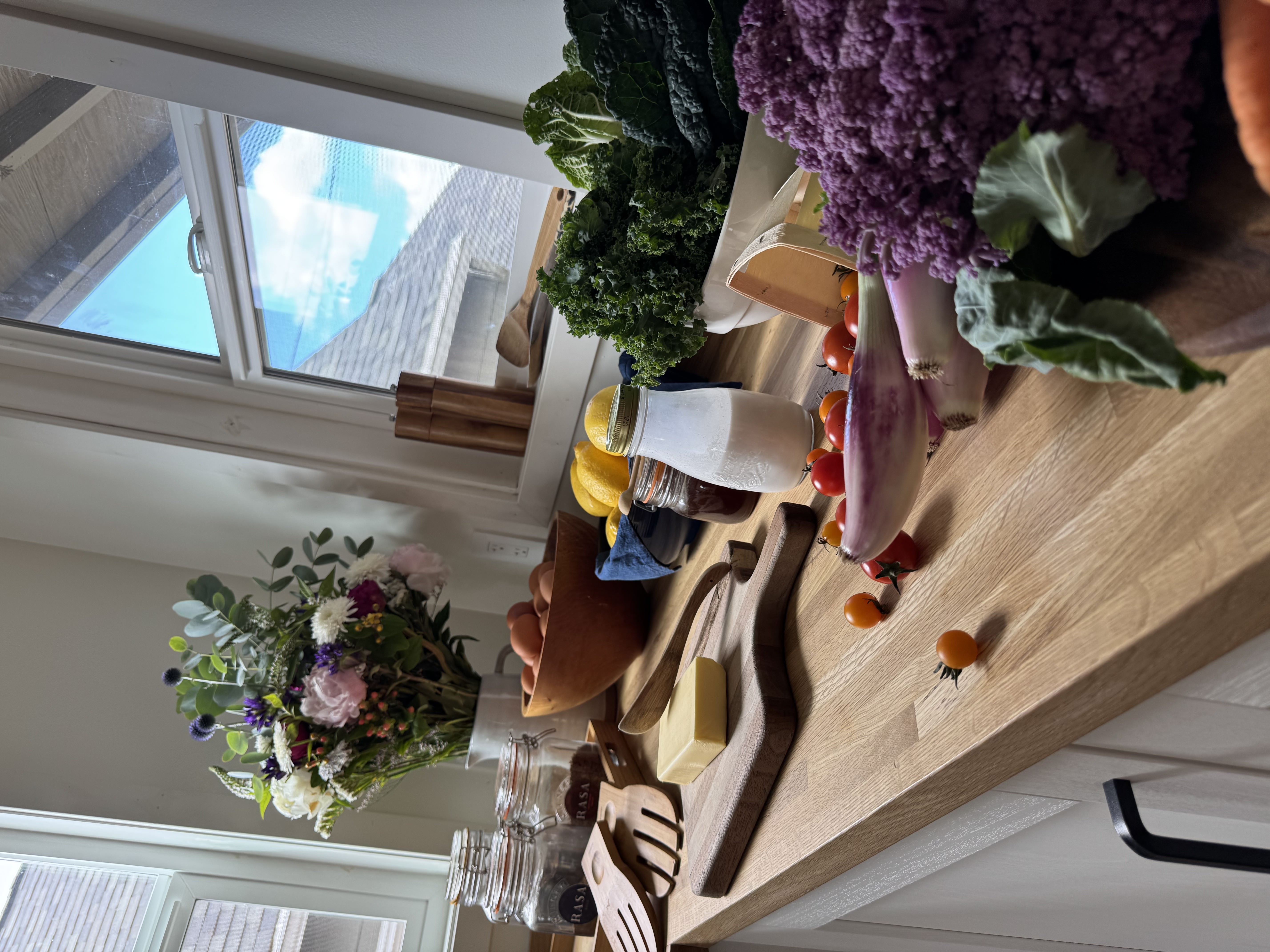 A kitchen counter with butter, eggs, lemons, milk, tomatoes and greens in window light