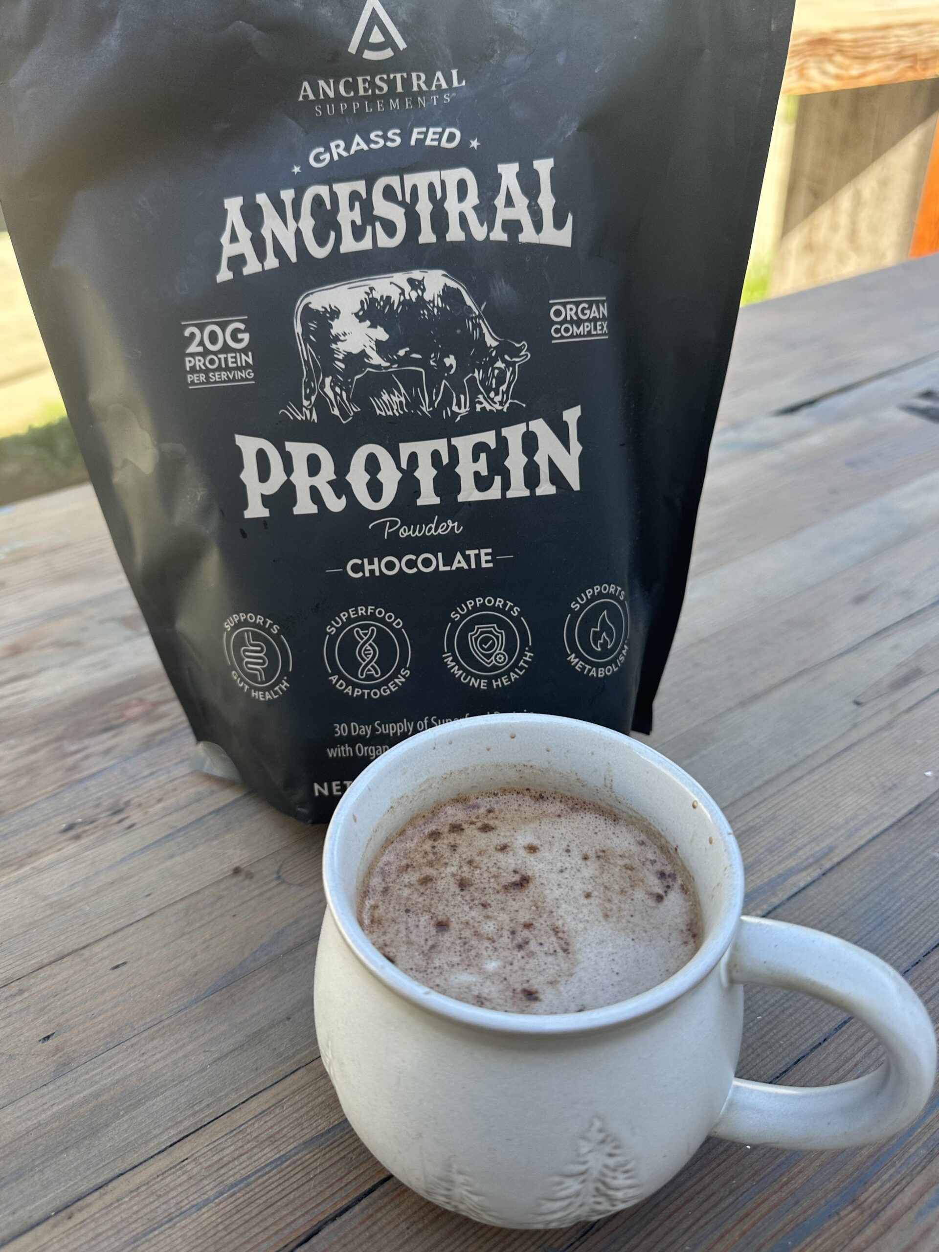 A frothy mug of dark protein cocoa on a wooden table in front of a bag of chocolate protein powder