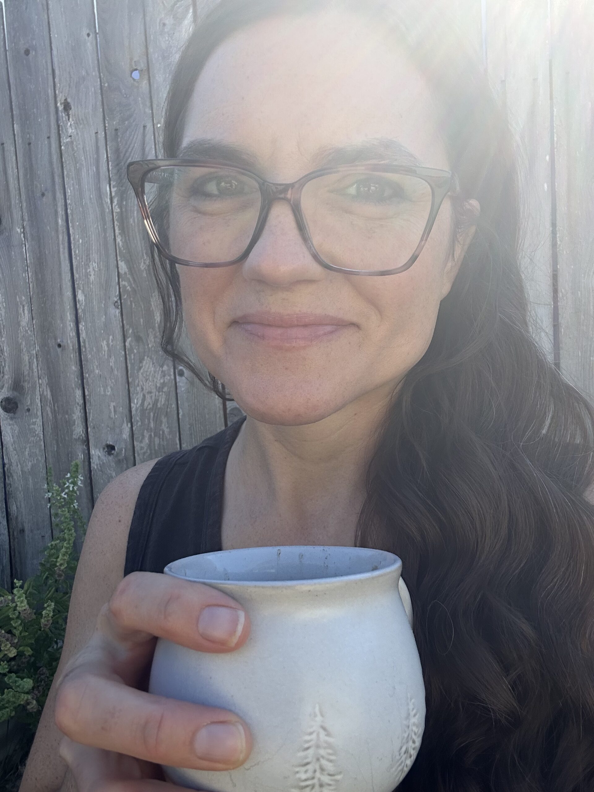 Brenna May holding a handmade ceramic mug of protein cocoa in front of a weathered wooden fence